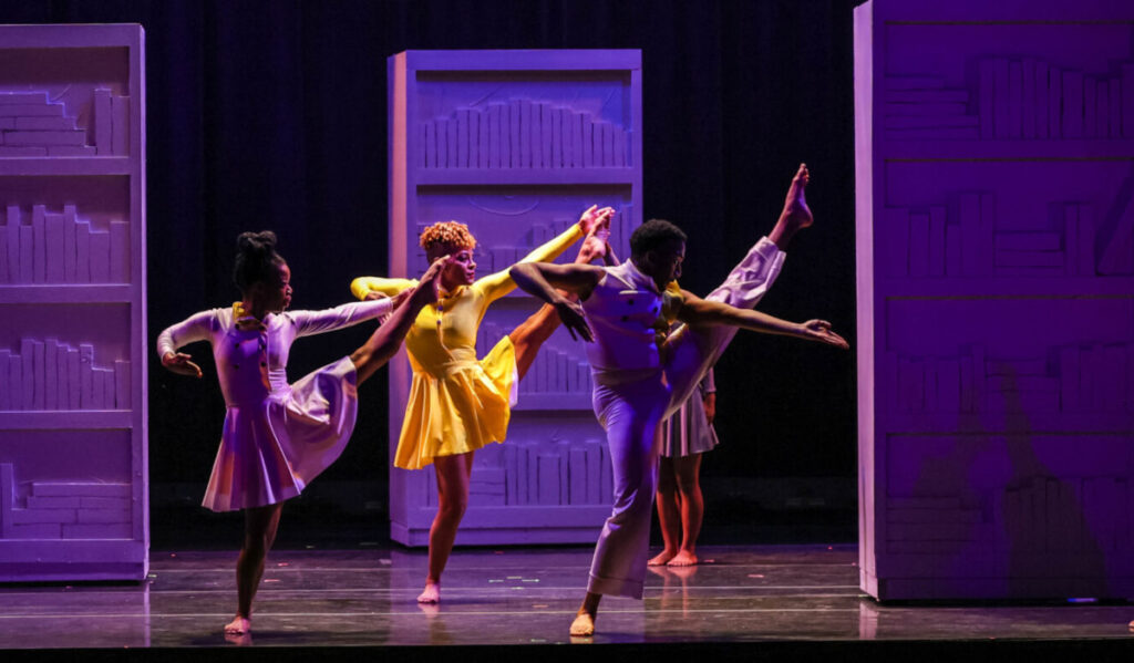 Three dancers on stage with a leg lifted high in purple and yellow light