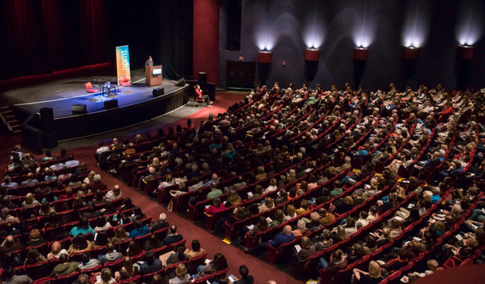 An auditorium with two empty chairs on stage and a crowd of people in seats