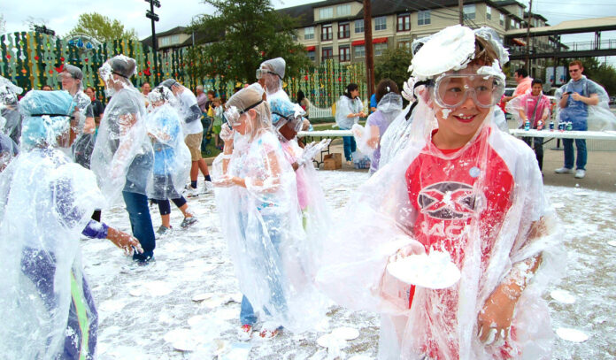 Children in plastic parkas are covered in shaving cream and holding plates to throw