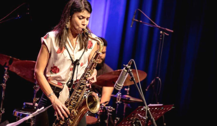 Melissa Aldana performing on a saxophone with a drummer behind her