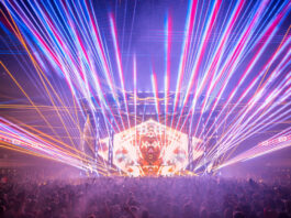 A concert stage covered in criss-crossed laser beams and the silhouette of a crowd in front of the stage