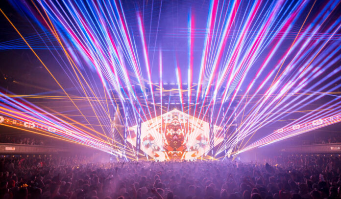 A concert stage covered in criss-crossed laser beams and the silhouette of a crowd in front of the stage