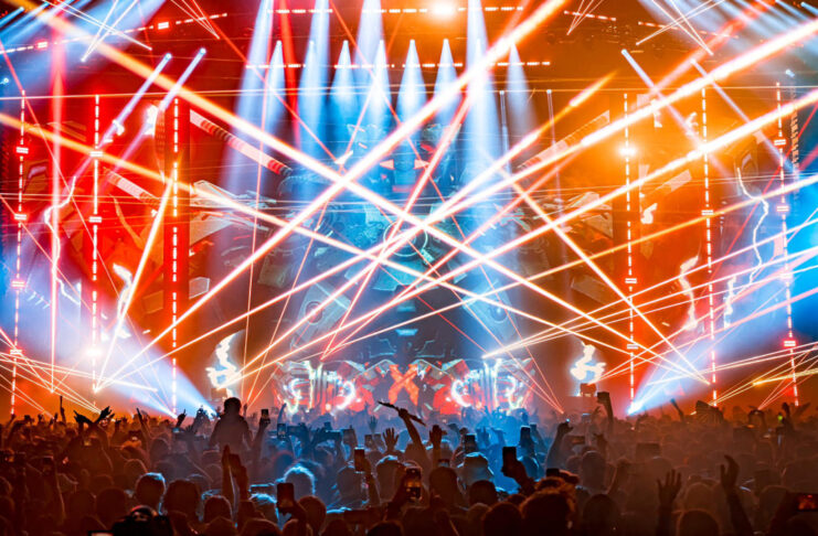 A concert stage covered in criss-crossed laser beams and the silhouette of a crowd in front of the stage