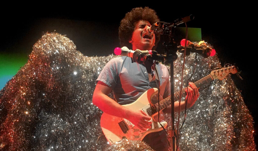 Helado Negro plays guitar and sings while people in tinsel costumes surround him