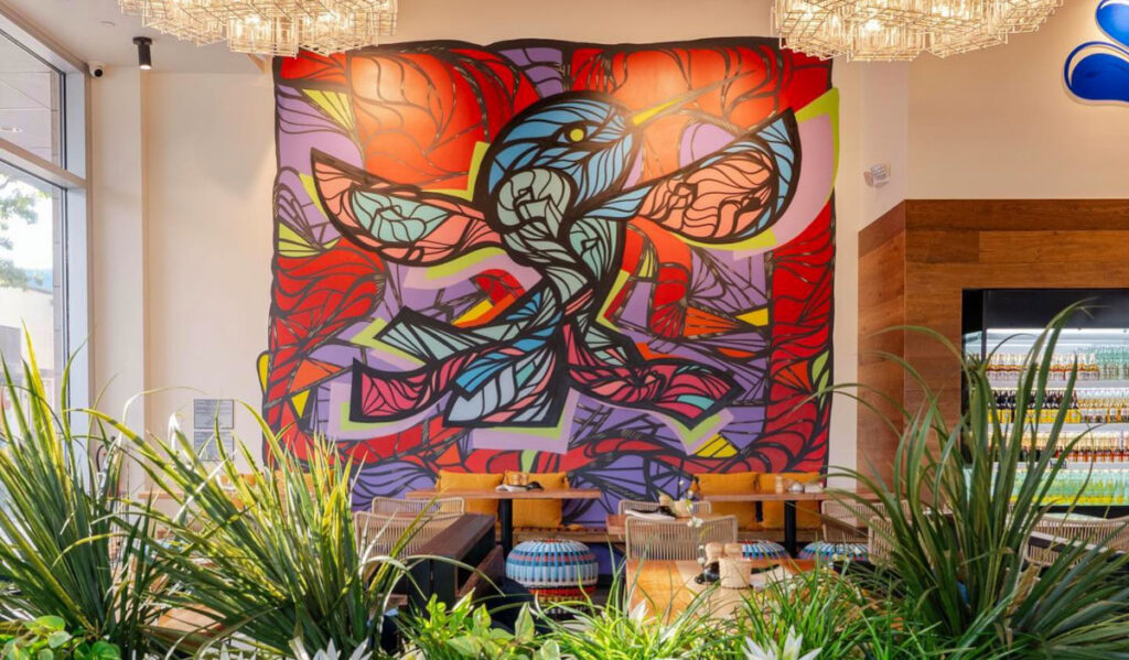 A dining room interior with leafy plants and a colorful abstract mural on the wall