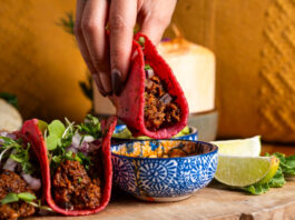A hand dipping a birria taco in a red tortilla into a sauce bowl