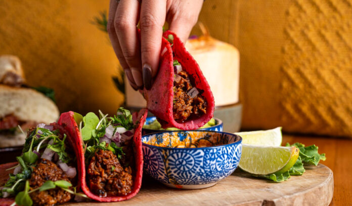 A hand dipping a birria taco in a red tortilla into a sauce bowl