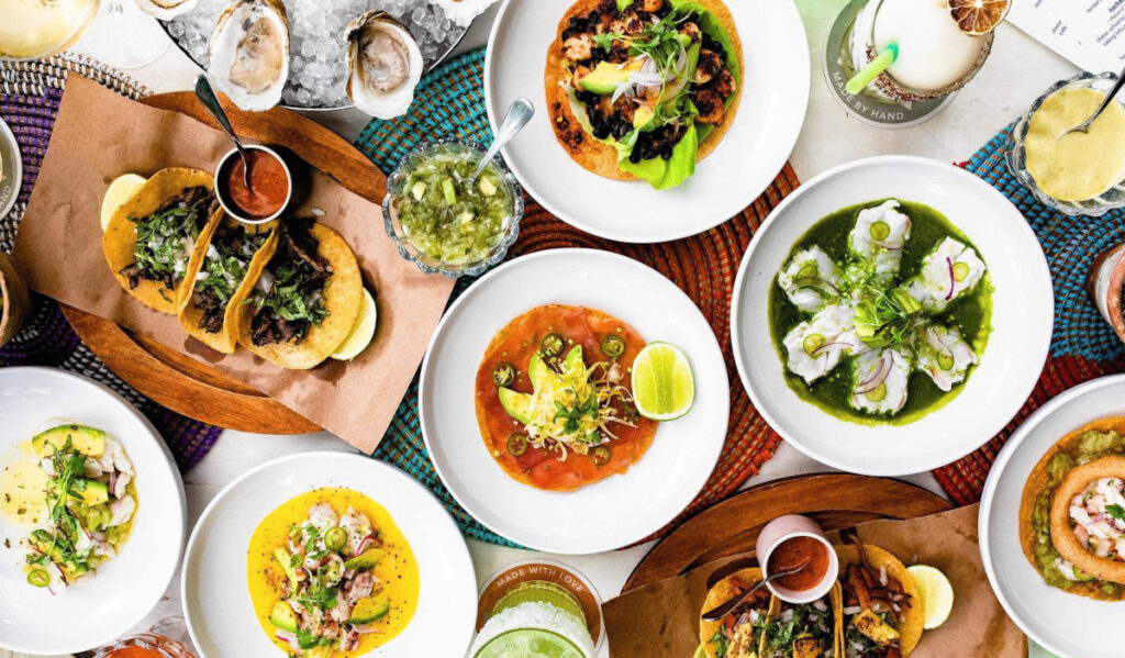 An overhead of multiple plates of dishes with tacos, soups, drinks and more