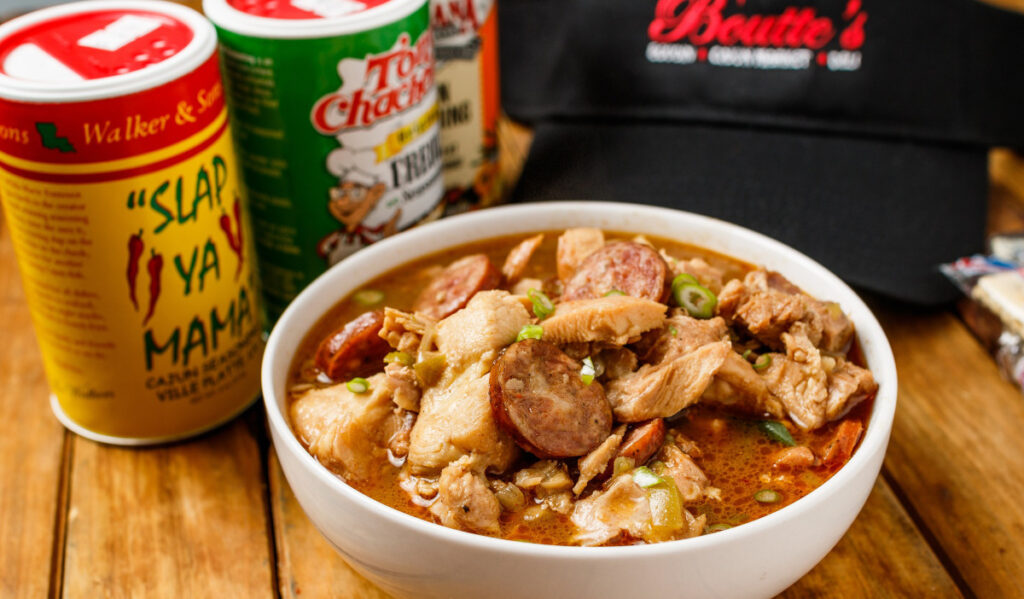 A bowl of gumbo next to two bottles of seasoning and a hat