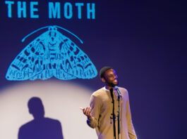 A performer on stage with "The Moth" projected behind them