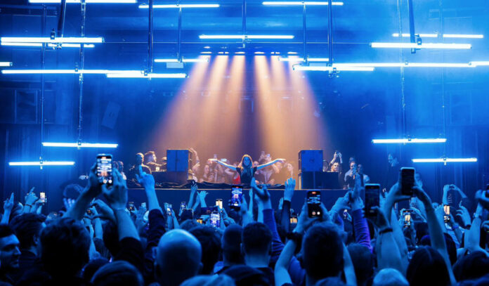 A DJ lit in gold light with arms oustretched in a blue-lit club