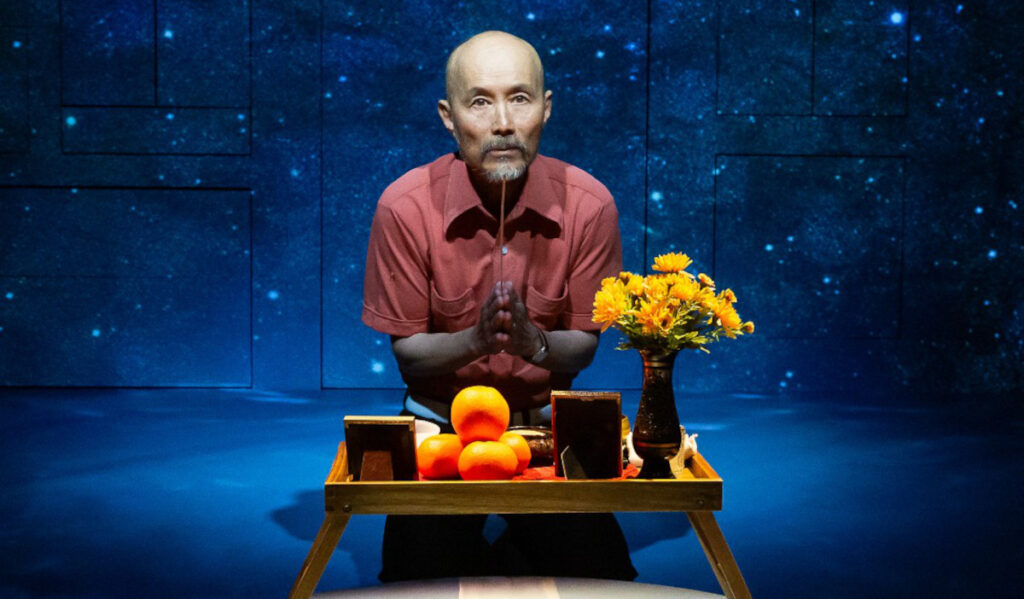 A man kneeling before a table with flowers, oranges and photos