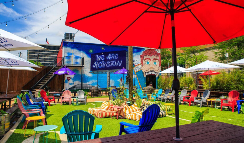 An outdoor beer garden on a sunny day with a "We Are Houston" mural