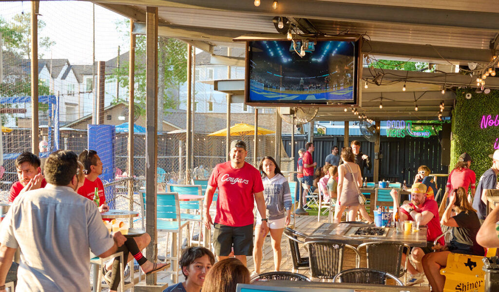 People walking through a covered patio area of a bar next to sand volleyball courts