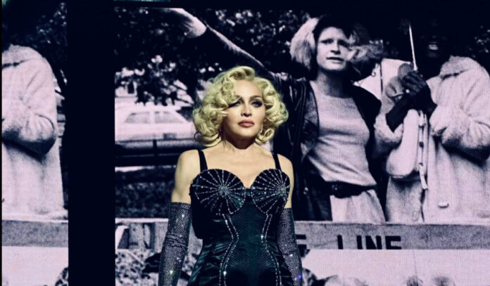 Madonna standing on stage with black and white images on a screen behind her