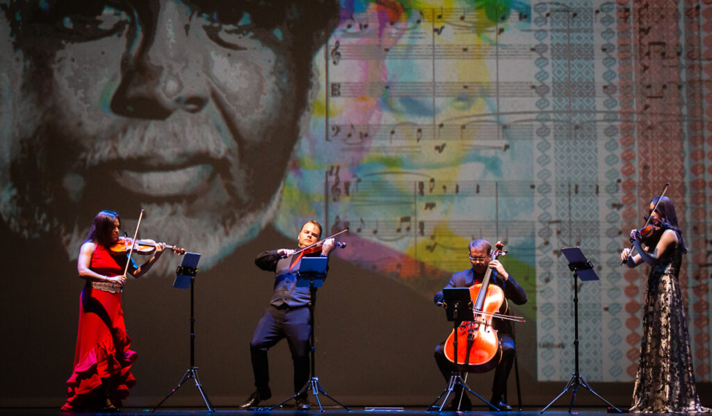 Three violinists and a cellist perform on stage with a projection of a face behind them