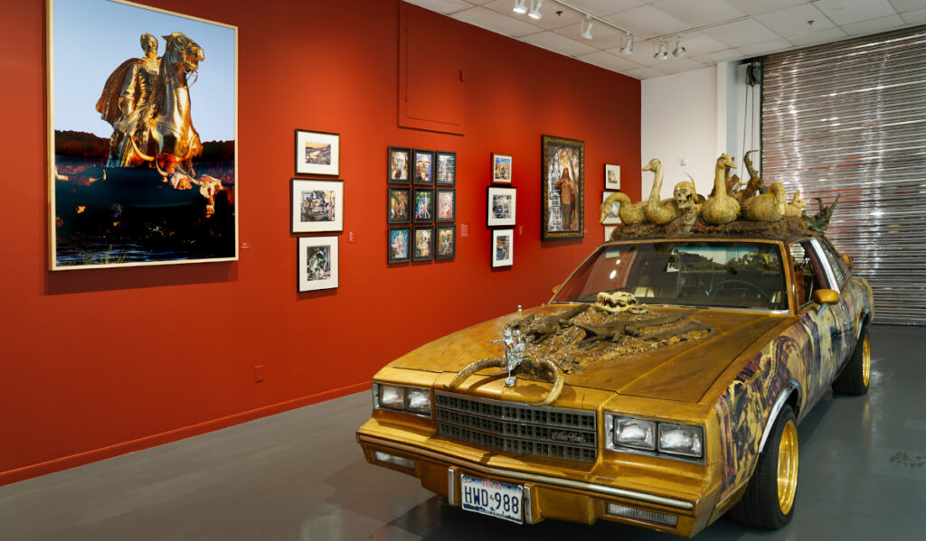 A golden car decorated with objects next to a red wall of collage artwork