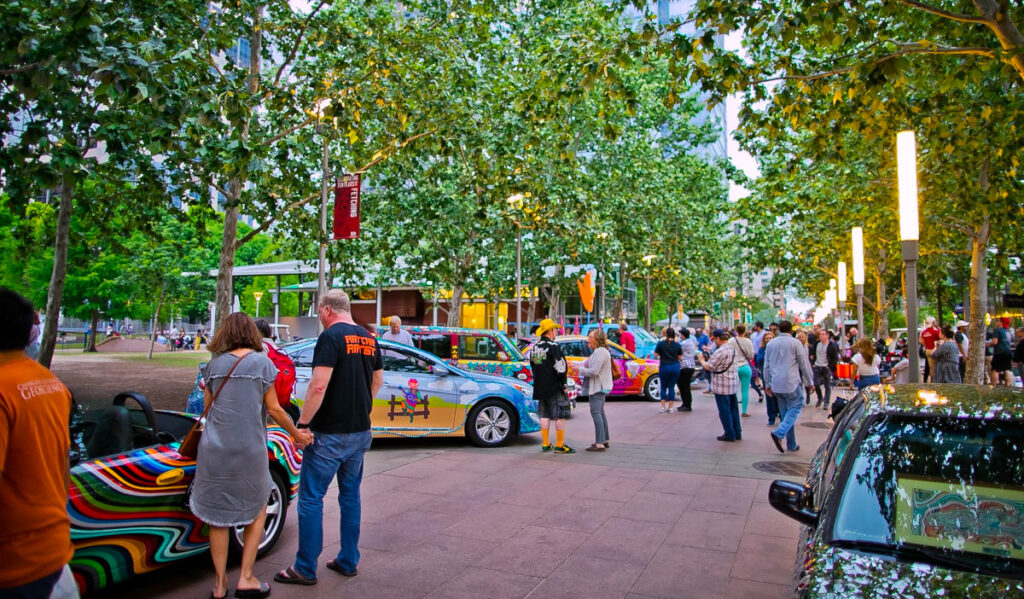 Art cars parked along a park path with people walking past and standing near them