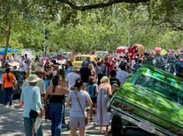 People walk around lowriders and art cars in a shaded street