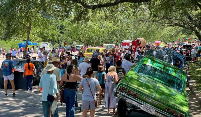 People walk around lowriders and art cars in a shaded street