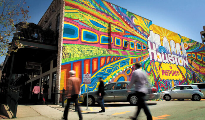 People walking by a vibrant and colorful mural on the side of a building that reads "Houston is Inspired"