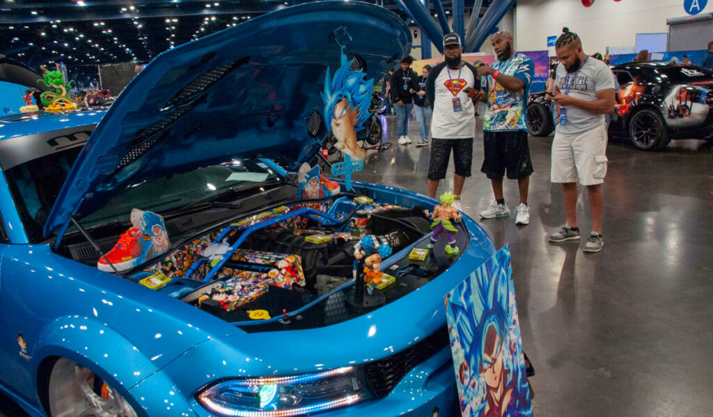 Three men standing next to a blue muscle car with a hood open and Dragonball Z decorations