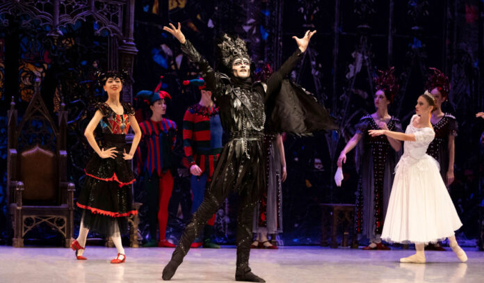 A performer dressed in black raises his arms as a ballerina in white and other actors stand behind