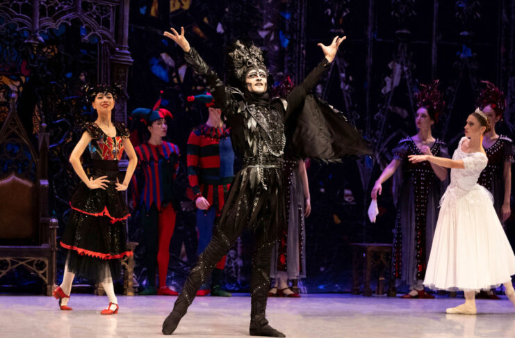 A performer dressed in black raises his arms as a ballerina in white and other actors stand behind
