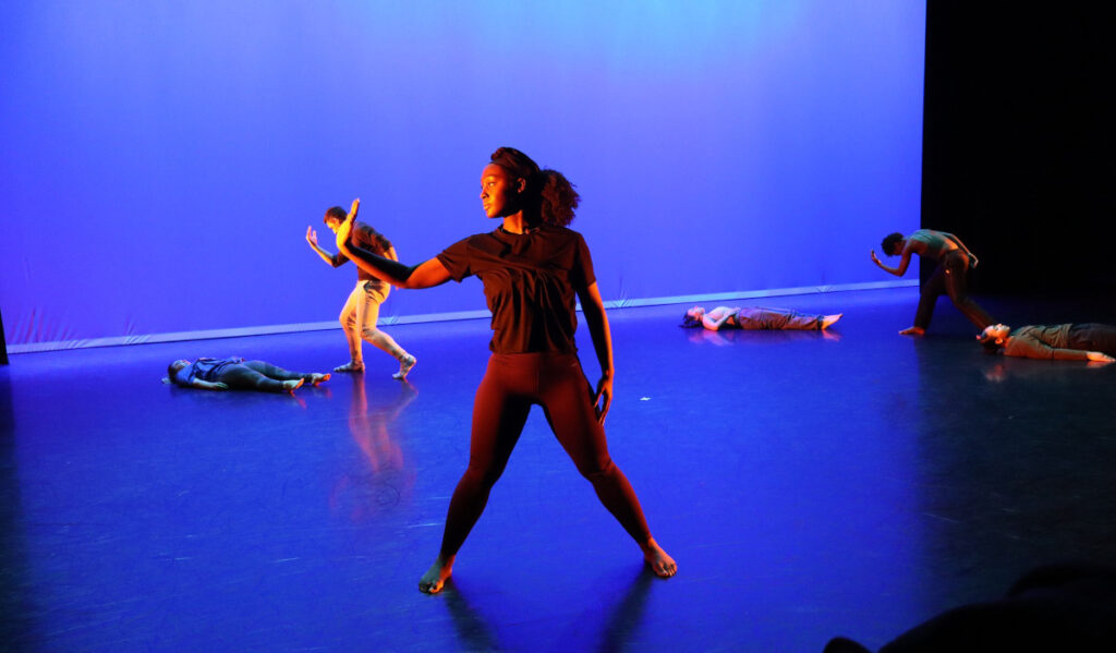 A women standing in front of a blue screen in pose, with other dancers behind her in various poses or laying