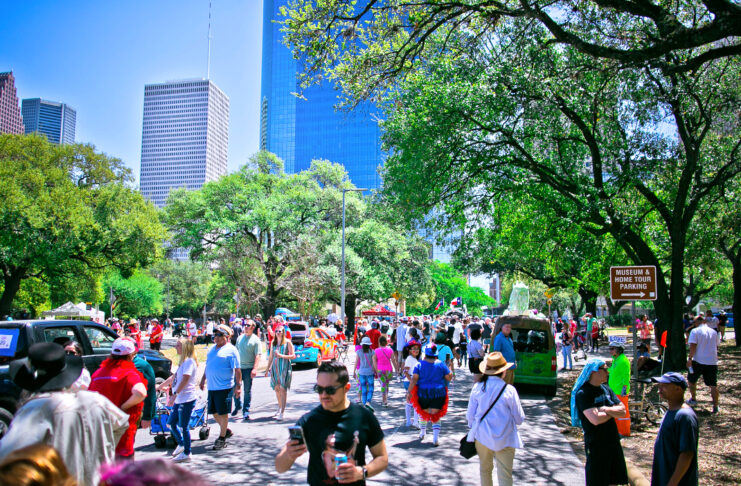 A bustling, sunny scene with people walking along streets past art cars and Downtown Houston in the background