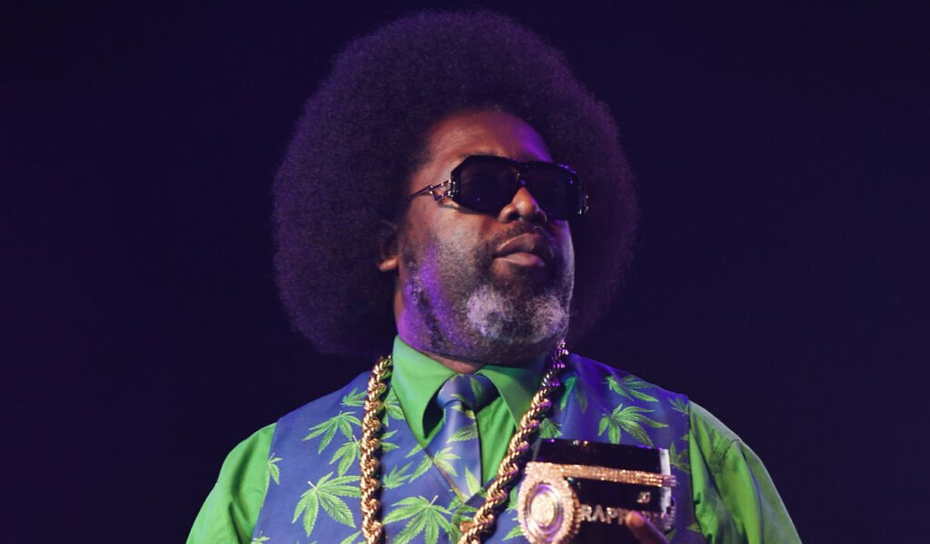 A close up of Afroman in a green and black suit with marijuana leaf pattern, holding a goblet