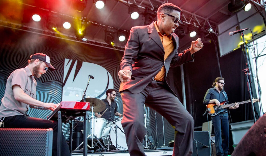 A singer wearing a suit and glasses dances on stage with his band behind him