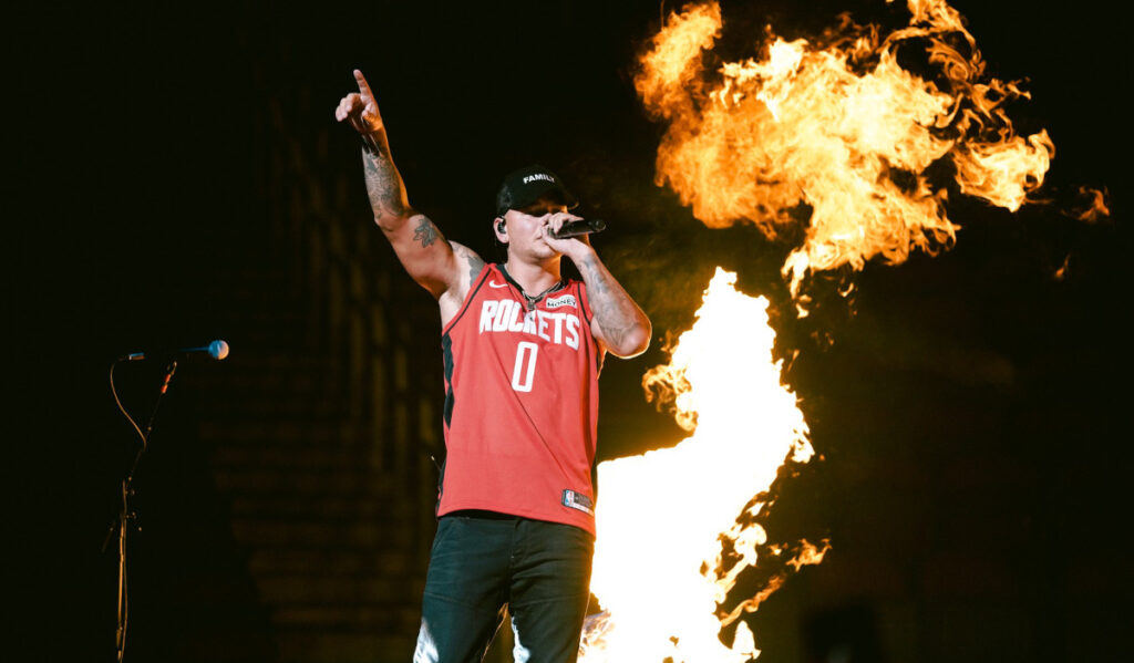 Kane Brown wearing a Rockets jersey lifting a hand to the crowd with a ball of flame behind him