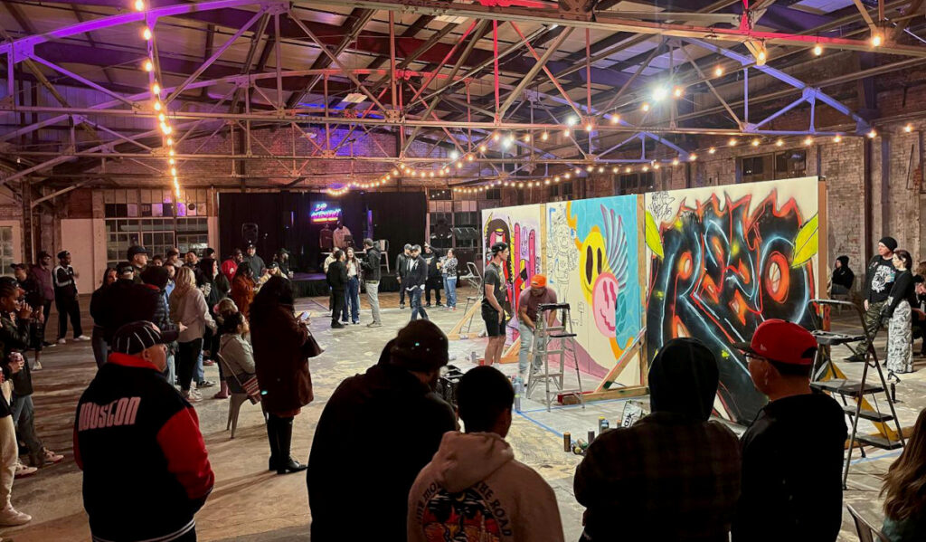 Artists paint graffiti on walls surrounded by a crowd of people