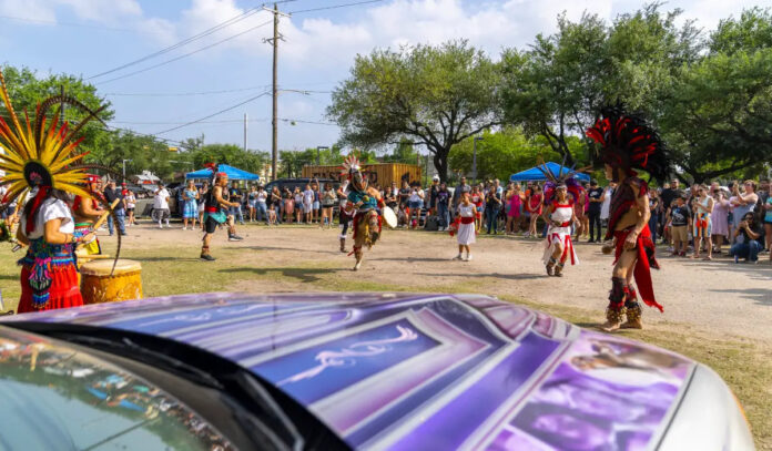 Indigenous dancers perform to a crowd next to lowriders