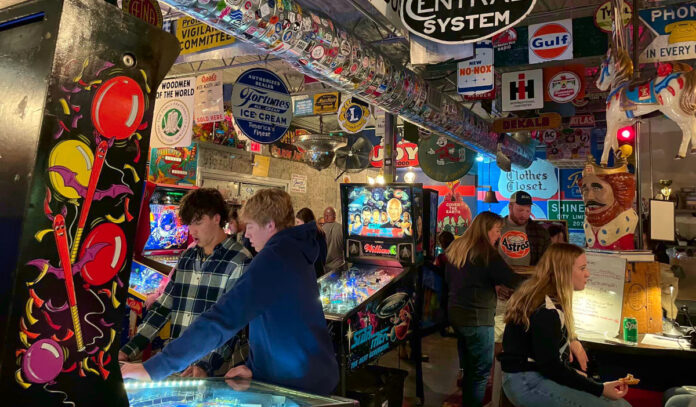 People stnading by pinball machines in a space decorated with hundreds of stickers