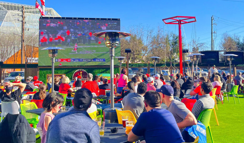 An outdoor terrace filled with groups of people watching sports on a giant screen