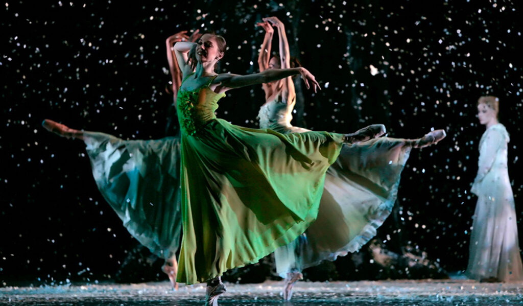 A ballerina in green balances on her leg as another ballerina behind her mirrors her