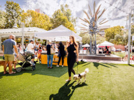 A woman walking a dog through a marketplace with customers at vendor booths