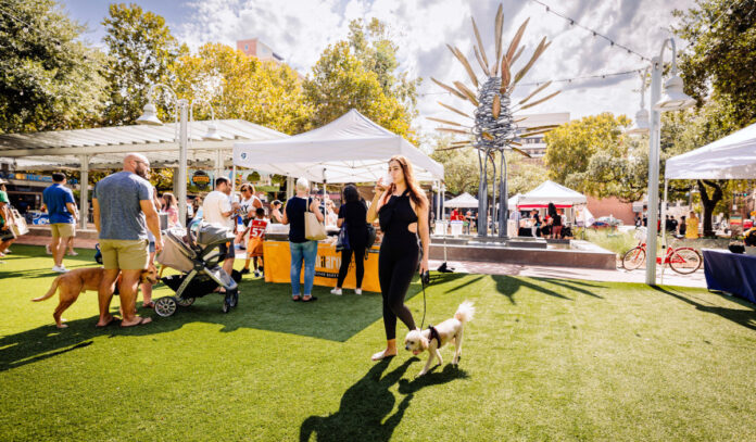 A woman walking a dog through a marketplace with customers at vendor booths