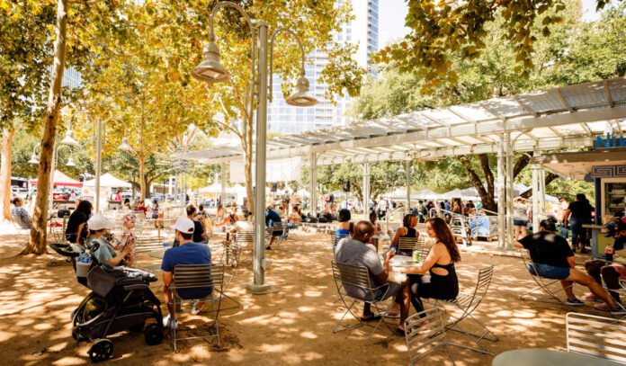 An outdoor scene under shady trees with people sitting at tables and shopping market stalls