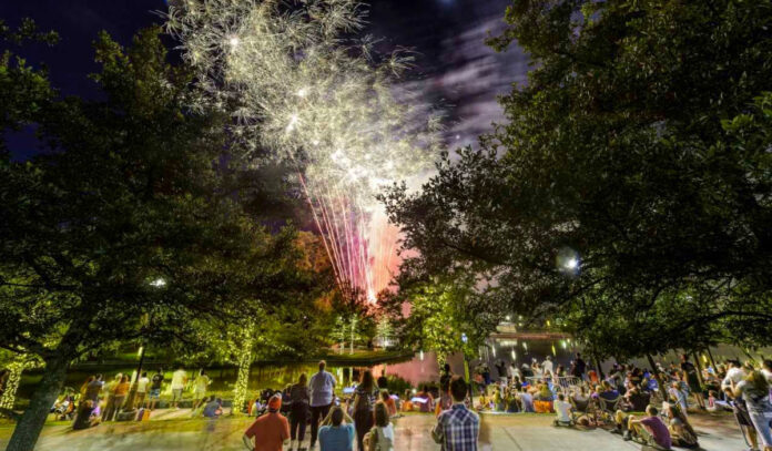 Red and white fireworks explode over a crowd of onlookers at a public park