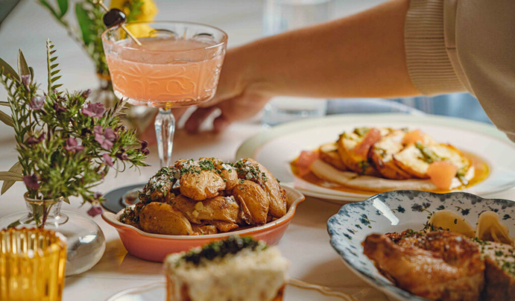 A hand outstretched behind a cocktail surrounded by plates of brunch food