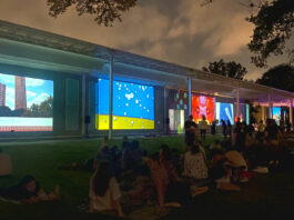 Crowds of people sit on a lawn as colorful projections are cast on the side of a museum