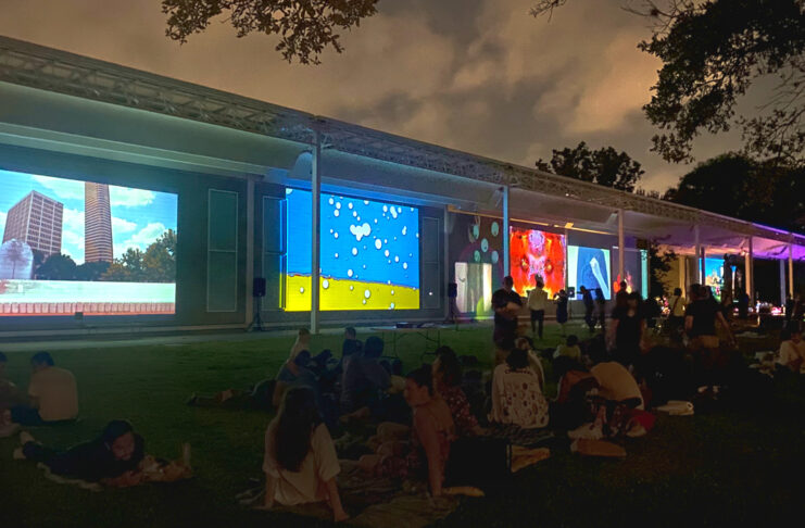 Crowds of people sit on a lawn as colorful projections are cast on the side of a museum