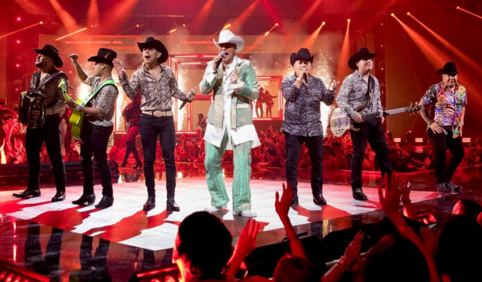 Seven members of Grupo Firme stand on a stage in a line with crowds around them