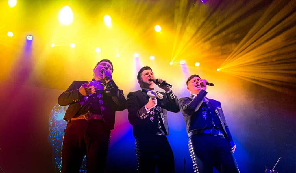 Three mariachi singers performing on stage with bright yellow and blue lights behind them