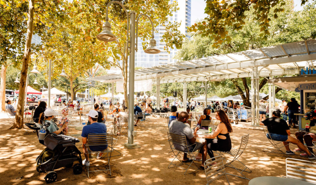 An outdoor scene under shady trees with people sitting at tables and market stalls