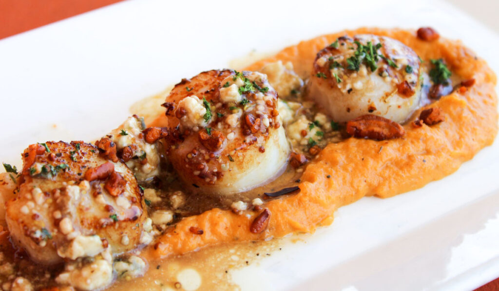 Three scallops on a plate in an orange sauce, topped with garnishes