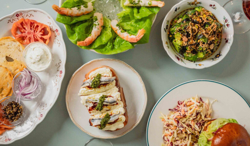 An overhead view of multiple plates of food, including shrimp tails, a burger and coleslaw, and toast